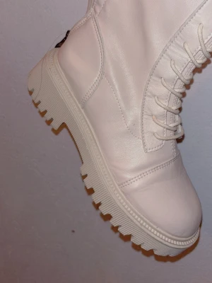 Vita höga chunky stövlar - Säljer ett par vita höga stövlar med chunky sula och snörning framtill. Stövlarna har dragkedja på insidan för enkel på- och avtagning. Tillverkade i ett slätt, matt material som liknar konstläder. Perfekta för dig som vill sticka ut med en trendig look.