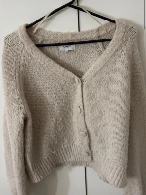 Beige kofta från ONLY med knappar - Mysig beige kofta från ONLY med v-ringning och tre matchande knappar framtill. Koftan är stickad i ett fluffigt material och har långa ärmar samt ribbade muddar vid ärmslut och nederkant. Perfekt för lager på lager och en chill look.