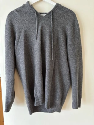 Mörkgrå Kashmir/merino hoodie - En stilren grå hoodie med huva och dragsnören. Tillverkad i mjuk ullmix som känns skön mot huden. Klassisk passform. Perfekt för en avslappnad look. LITET HÅL VID EN ARMBÅGE MEN ENKELT ATT LAGA/DÖLJA.