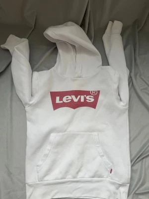 Vit hoodie från Levi's med logga - Säljer en vit hoodie från Levi's med stor röd logga på bröstet. Hoodien har en klassisk känguruficka framtill och mjukt, fleeceliknande foder på insidan. Perfekt för dig som gillar streetwear och vill ha något bekvämt och stilrent.   Passar även 11 år den lilla minimal pricken går inte bort den syns knappt om man tvättat odenkligt och har typ nåt medel då kan den gå bort den är minimal!!! Skriv om du är intresserad eller om du har några frågor. Perfekt nu för vintren 