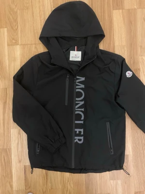 Svart vindjacka från Moncler - Snygg svart vindjacka från Moncler med stor logga tryckt vertikalt på framsidan. Jackan har huva, två dragkedjeförsedda fickor och en bröstficka. Elastiska muddar vid ärmslut och justerbar nederkant med dragsko. Perfekt för dig som gillar streetwear och exklusiva märken. Den är storlek  M, men passar också på S men lite storre.