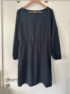 Mörkblå Åhléns-klänning, storlek 42 - Stilren mörkblå / marinblå klänning från Åhléns i storlek 42. Klassisk rundad halsringning och långa ärmar med lätt puffärm och dekorativa stockning i ärmsluten. Diskret och praktisk resår i midjan för en bekväm passform. Stängs med en fin klädd knapp i nacken. Supersnygg som den är eller med skärp i midjan. Bra modell som lätt går att både styla upp för fest eller styla ned för jobb/vardag. Mycket fint skick. 