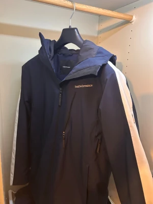 Mörkblå vindjacka Peak Performance - Snygg mörkblå vindjacka från Peak Performance med vit logga på bröstet och stor vit detalj på huvan. Perfket för skidåkning ny pris:3499kr