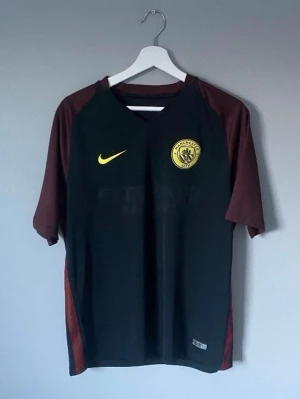Manchester City matchtröja Nike - Snygg Manchester City matchtröja från Nike i svart med vinröda ärmar och gula detaljer. Tröjan har klubbmärke på bröstet och Nike-logga. Perfekt för fotbollsträning eller att bära till vardags för fans. Vet inte vad de är för storlek men skulle säga L
