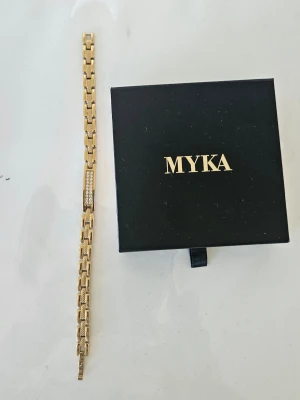 Guldfärgat armband från MYKA - Snyggt guldfärgat armband från MYKA med rektangulära länkar och en detalj med gnistrande stenar på ovansidan. Armbandet har en elegant och modern design som passar perfekt till din stil. Levereras i original ask. Armbandet är ca 21 cm långt.