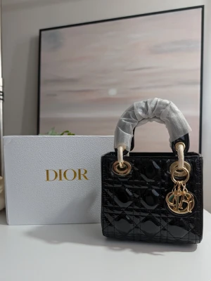 Lady DlOR väska Mini ♥️ - Denna exklusiva handväska är en symbol för tidlös sofistikering. Med sin högblanka finish och sitt karaktäristiska geometriska sömnadsmönster är den det ultimata valet för den som söker en klassisk men ändå modern accessoar.En oerhört chic och glamorös väska som utstrålar självsäkerhet. Den är perfekt för såväl viktiga affärsmöten som eleganta kvällsevenemang.