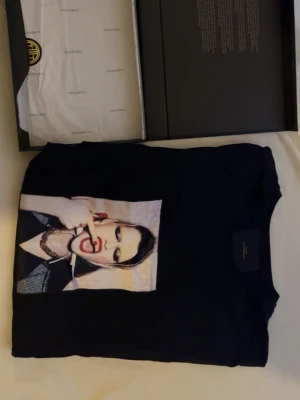 Svart t-shirt Lanvin med grafiskt tryck - Cool svart t-shirt från Limitato med ett stort grafiskt tryck på bröstet som föreställer en person med smink och smycken. T-shirten har klassisk passform och är gjord i mjuk bomull. Perfekt för dig som vill sticka ut med en unik design.