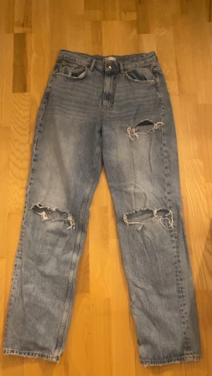 Blåa ripped jeans - Säljer ett par ljusblå baggy jeans med flera slitningar och hål på både fram- och baksida. Jeansen har raka ben och klassiska fem fickor. Perfekta för en avslappnad och trendig look.