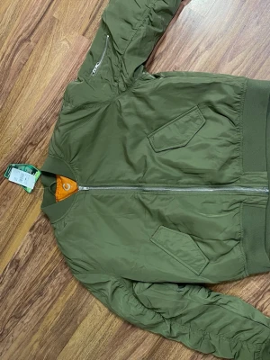 Olivgrön bomberjacka med orange foder - Snygg olivgrön bomberjacka med klassiskt orange foder och silverfärgad dragkedja. Jackan har två snedställda fickor framtill och en ärmficka med dragkedja. Ribbstickade muddar vid ärmslut och midja. Perfekt för en cool streetstil.