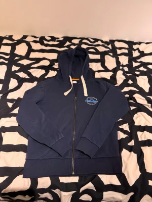 Mörkblå zip hoodie från jack&jones - Mörkblå hoodie från Jack & Jones med dragkedja framtill och vit texttryck på bröstet. Huva med vita snören och ribbade muddar vid ärmslut och nederkant.