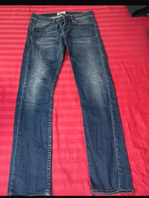 Blå raka jeans med slitningar - Snygga blå jeans med raka ben och diskreta slitningar på låren. Klassisk femficksmodell med normal midja och dragkedjegylf. Jeansen har en tidlös tvätt och passar perfekt till en avslappnad stil.