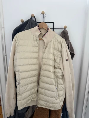Beige pufferjacka från Moncler - Stilren beige pufferjacka från Moncler med quiltad front och släta ärmar. Jackan har ståkrage med röd, vit och blå rand, två fickor med tryckknappar och Moncler-logga på ärmen. Perfekt för kalla vinterdagar och ger en sportig vibe.