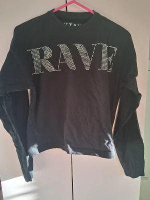 Svart långärmad topp med text - Svart långärmad topp med stora bokstäver 'RAVE' framtill i glittriga stenar. Rund halsringning och avslappnad passform. Perfekt för dig som gillar statement-plagg och vill sticka ut. Det har ramlat av några pärlor därför jag säljer den billigt