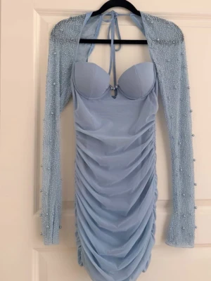 Ljusblå ruched klänning med pärlor - Supersnygg ljusblå kortklänning med ruched tyg och vadderad byst. Klänningen har avtagbara långa ärmar dekorerade med pärlor och knytning i nacken. Perfekt för dig som vill sticka ut med en unik och trendig look.