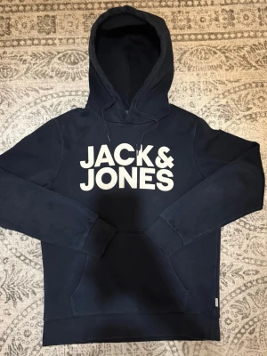 Mörkblå hoodie från Jack & Jones - Snygg mörkblå hoodie från Jack & Jones med stor vit logga framtill. Klassisk modell med huva och dragsko samt en stor magficka. Perfekt för en avslappnad och trendig stil. Mjuk insida för extra komfort.