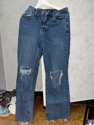 Blå slitna raka jeans med hål st 36 - Säljer ett par blå jeans med raka ben och stora slitna hål på båda knäna. Jeansen har klassiska fickor och råa kanter nertill. Perfekta för en avslappnad och trendig streetstil.