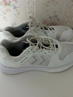 Vita sneakers från Hummel Power Play Pro - Snygga vita sneakers från Hummel Power Play Pro med klassisk snörning och meshdetaljer på sidorna. Skorna har en sportig design med Hummels ikoniska pilar och en stabil sula som passar perfekt för en aktiv livsstil.