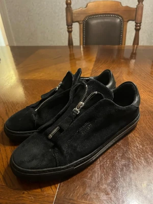 Svarta sneakers från Axel Arigato - Snygga svarta sneakers från Axel Arigato i mocka med coola dragkedjor istället för vanliga snören. Låg modell med svart sula och diskret logga på sidan. Perfekta för dig som gillar stilrena och unika detaljer på dina skor.