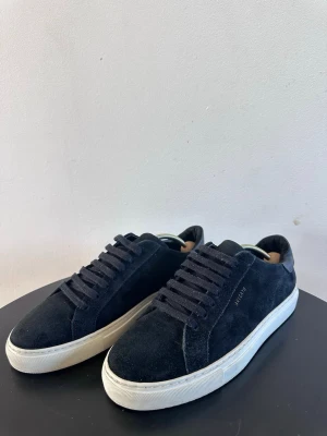 Svarta sneakers från Axel Arigato - Stilrena svarta sneakers i mocka från Axel Arigato med vita sulor och svarta skosnören. Klassisk låg modell med diskret logga på sidan. Perfekta för en clean och modern look.