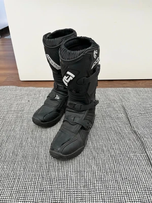 Svarta crossstövlar från Moose Racing - Säljer ett par svarta crossstövlar från Moose Racing med kraftiga spännen och förstärkta skydd vid smalben och fot. Stövlarna har en sportig look och är tillverkade i slitstarkt syntetmaterial och plastdetaljer. Perfekta för dig som gillar motocross eller enduro.