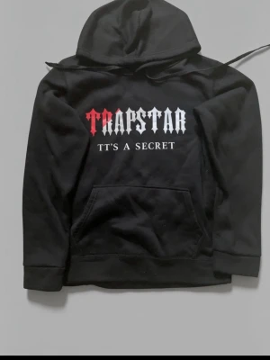 Svart Trapstar hoodie med tryck - Svart hoodie från Trapstar med huva och stor ficka framtill. Framsidan har ett tryck i rött och vitt med texten 'TRAPSTAR IT'S A SECRET'. Perfekt för en streetwear-look och riktigt skön att bära.