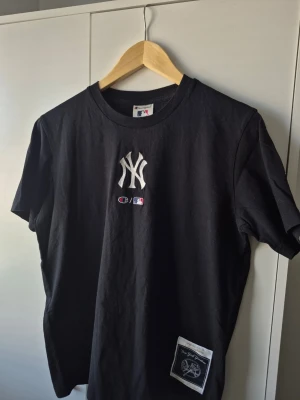 New York Yankees x Champion - Svart t-shirt från Champion med New York Yankees-logga broderad på bröstet och patch nertill. Klassisk rund hals och korta ärmar. Perfekt för dig som gillar sportig streetstyle och vill sticka ut med en ikonisk MLB-look.