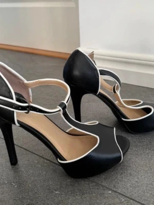 Svarta pumps med vit kant - Snygga svarta pumps från H&M med vit kontrastkant och T-rem. Skorna har öppen tå och hög klack, vilket ger en elegant och trendig look. Tillverkade i konstläder med justerbart spänne vid ankeln.