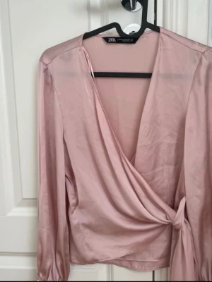 Rosa omlottblus från Zara - Snygg ljusrosa omlottblus från Zara i glansigt satinliknande material. Djup v-ringning och knytning i sidan ger en trendig look. Långa ärmar med lätt puff vid axlarna och smala muddar vid ärmslut. Perfekt för en chic och modern stil.