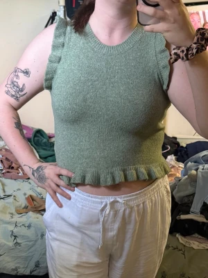Grön stickad ärmlös topp med volang - Superfin ljusgrön stickad topp med volangdetaljer vid ärmöppningarna och nederkanten. Modellen är croppad och har en rund halsringning. Perfekt att styla med höga byxor eller kjol för en trendig look.