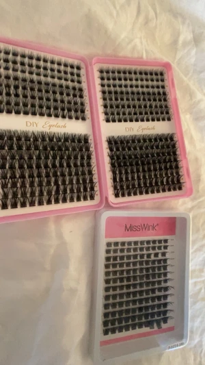 DIY och MissWink lösögonfransar kit - Säljer flera kit med svarta individuella ögonfransar från MissWink och DIY Eyelash. Fransarna kommer i olika längder och tjocklekar, perfekta för att skapa en personlig look. Förpackningarna är rosa och vita och fransarna är gjorda av syntetmaterial.