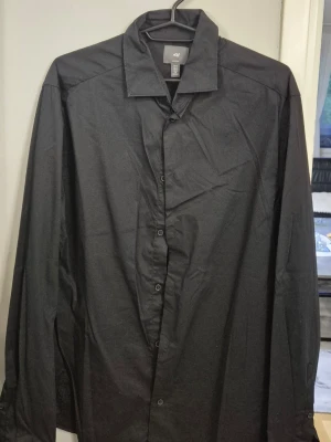 Svart slim fit skjorta från H&M - Säljer en svart långärmad skjorta från H&M i slim fit-modell. Skjortan har klassisk krage, knäppning framtill och är tillverkad i ett tunt, mjukt tyg som ger en clean look. Perfekt för dig som gillar stilrena och enkla plagg.