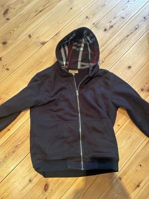 Svart Burberry hoodie med dragkedja - Snygg svart hoodie från Burberry med dragkedja framtill och klassiskt rutigt foder i huvan. Hoodien har känguruficka och ribbade muddar. Perfekt för dig som vill ha en stilren och exklusiv look med ikoniska Burberry-detaljer.