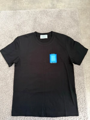 Svart t-shirt från Casablanca - Svart t-shirt från Casablanca med blått grafiskt tryck och texten 'Casa Blanca' på ryggen. Klassisk rund hals och korta ärmar. Perfekt för dig som gillar stilrena plagg med en twist.