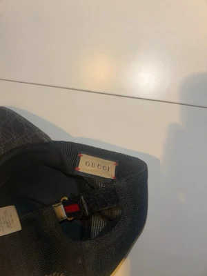 Svart keps från Gucci med monogram - Snygg svart keps från Gucci med klassiskt GG-monogram på skärmen och röd-blå rand på sidan. Kepsen har justerbar rem baktill och är tillverkad i ett slitstarkt tygmaterial. Perfekt accessoar för att lyfta din streetwear-look.