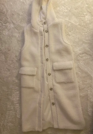 Vit lång teddyväst med huva - Vit lång teddyväst med huva och stora fickor framtill. Mjuk och varm modell med knäppning framtill.  Helt oanvänd.