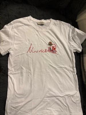 Vit Moncler t-shirt med röd logga - Snygg vit t-shirt från Moncler med röd broderad logga och text på bröstet. Klassisk passform och rund halsringning. Perfekt för en clean och stilren look.