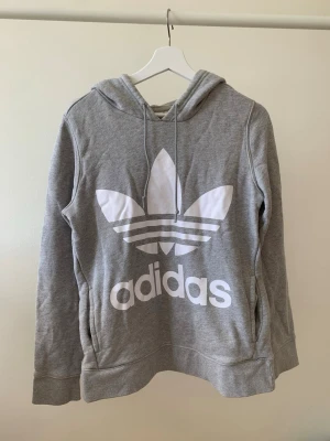 Grå Adidas hoodie med stor logga - Snygg grå hoodie från Adidas med stor vit Trefoil-logga och text på bröstet. Mjuk insida, dragsko i huvan och känguruficka framtill. Perfekt för en avslappnad streetstyle-look. Tillverkad i bomullsblandning.