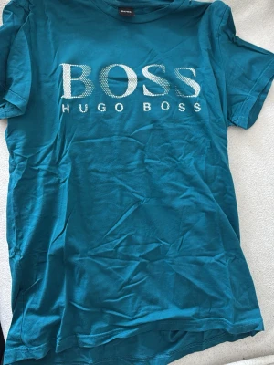 Turkos Hugo Boss t-shirt med tryck - Snygg turkos t-shirt från Hugo Boss med stort vitt logotyptryck på bröstet. Klassisk rund halsringning och korta ärmar. Perfekt för dig som vill ha en stilren och trendig look med en touch av premiumkänsla.