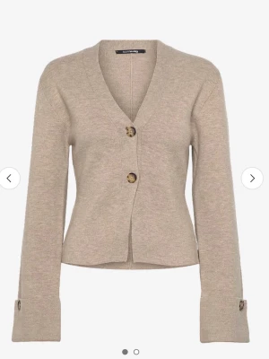 Beige kofta från Gina  - Stilren beige kofta med v-ringning och två stora knappar framtill. Koftan har långa ärmar med dekorativa knappar vid ärmsluten och är tillverkad i ett mjukt stickat material. Perfekt för lager-på-lager och enkel att matcha med olika outfits. Knappt använd, nypris 500