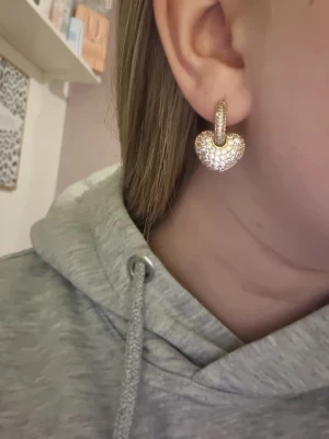 Guldiga hjärtörhängen med stenar - Snygga örhängen i guldton med hjärtformad design, täckta av glittrande stenar. De har en modern och trendig look som verkligen sticker ut. Perfekta för att ge din outfit lite extra bling och romantisk vibe.