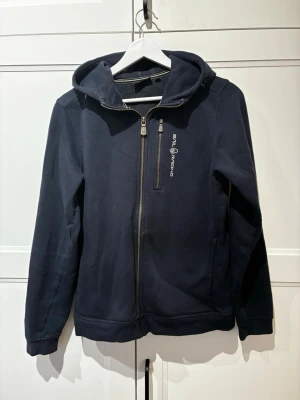 Marinblå hoodie från Sail Racing - Snygg marinblå hoodie från Sail Racing med dragkedja hela vägen och en extra bröstficka med dragkedja. Hoodien har en klassisk huva och logga tryckt på bröstet. Perfekt för dig som gillar sportig och stilren look. Nypris (999)
