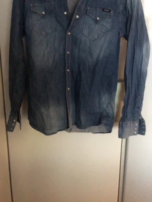 Blå jeansjacka från Replay - Snygg blå jeansjacka från Replay med klassisk krage, tryckknappar och två bröstfickor med lock. Jackan har långa ärmar med knappslå vid ärmslut och är tillverkad i mjukt denimtyg. Perfekt för en avslappnad och trendig look.