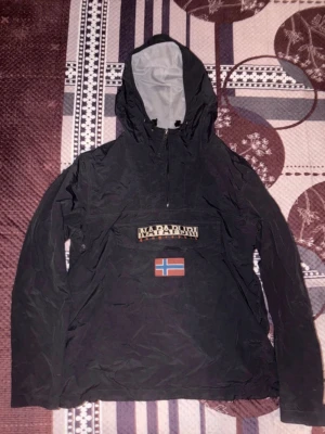 Svart vindjacka från Napapijri - Äkta Svart vindjacka från Napapijri med huva och halv dragkedja. Originellt köpt från zalando 2495kr men säljer för 800kr eftersom den är för liten och lite använd! Stor logotyp på bröstet samt norsk flagga framtill. Jackan har en sportig look och är tillverkad i ett lätt, vindtätt material. Perfekt för blåsiga dagar.