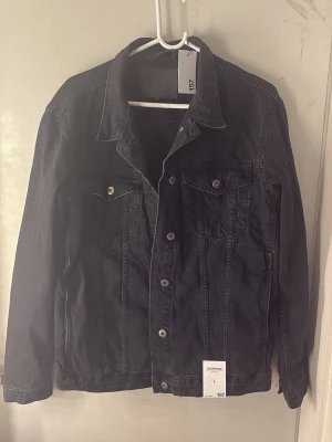 Svart jeansjacka  - Snygg svart jeansjacka med klassisk krage, knäppning framtill och två bröstfickor med lock och knapp. Jackan har raka ärmar och en stilren look som passar till det mesta. Perfekt för dig som gillar en enkel men trendig stil. Helt ny oanvänd, prislapp kvar! Köptes för 400 kr