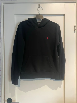 Svart hoodie från Ralph Lauren - Svart hoodie från Ralph Lauren med klassisk huva och känguruficka. Liten röd broderad logga på bröstet. Mjuk och skön bomullskänsla, perfekt för en avslappnad stil. Långa ärmar och ribbade muddar för extra komfort.