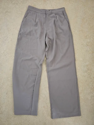 Grå kostymbyxor med raka ben - Snygga grå kostymbyxor med hög midja och raka ben. Byxorna har pressveck framtill, sidfickor och en bakficka. Perfekta för en clean och stilren look. Materialet känns mjukt och har en lätt glans.