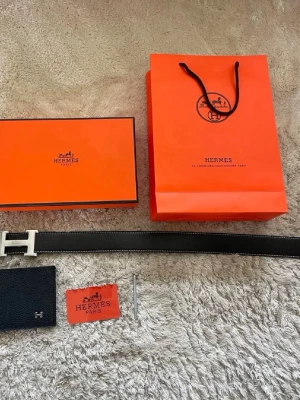 Svart skärp med H spänne - Säljer ett svart skärp i skinn med ett H-spänne i silver. Bältet har vit kontrastsöm och kommer med original förpackning, påse och certifikat. Perfekt accessoar för att lyfta vilken outfit som helst. Bältet är 110 cm och priset kan diskuteras. Det är bara att skriva vid minsta intresse