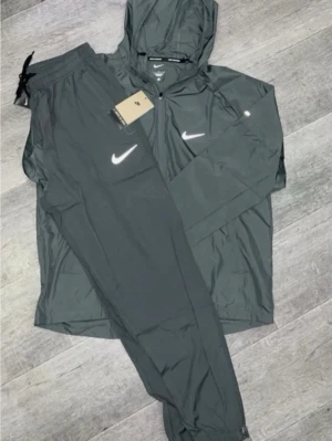 Nike miller tracksuit - Snyggt svart träningsset från Nike med matchande jacka och byxor. Jackan har huva, dragkedja och vita Nike-loggor på bröstet och byxan. Perfekt för träning eller chill. Lätt och smidigt material som andas.