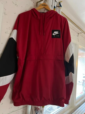 Röd Nike Air vindjacka med huva - Röd vindjacka från Nike Air med svart och vitt mönster på ärmarna. Jackan har huva, halv dragkedja framtill och elastiska muddar. Stor Nike Air-logga på bröstet. Perfekt för en sportig och avslappnad stil.