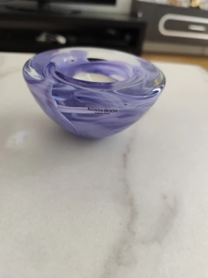 Lila glaslykta från Kosta Boda - Säljer en snygg lila glaslykta från Kosta Boda med virvlande mönster. Den är rund och har en skålformad design, perfekt som ljushållare eller dekoration. Tillverkad i glas och har en modern vibe som passar i de flesta rum.